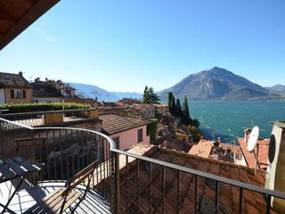 Panoramic view from a balcony over terracotta roofs towards the calm waters of Lake Como and a mountainous horizon under a clear sky.