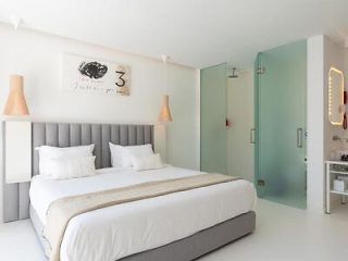 Modern hotel room with a large bed featuring a gray headboard, flanked by warm-toned lamps, with minimalist decor and an open bathroom concept