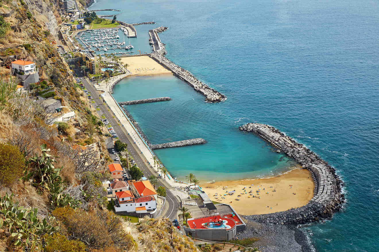 An aerial view of a beachside town with a marina, protected swimming area, and a curving shoreline against a backdrop of hills