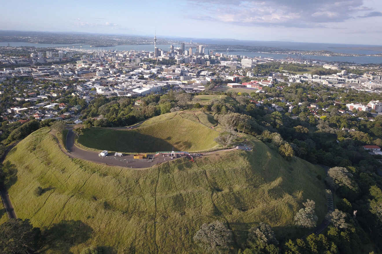 An aerial view of a lush, grass-covered volcanic cone in a city park, with a panoramic backdrop of a sprawling cityscape and harbor