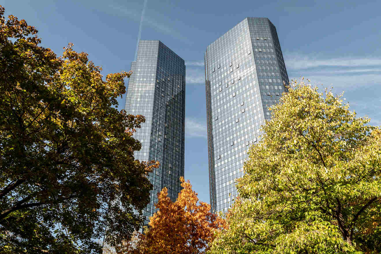 Autumnal trees framing the towering Deutsche Bank Twin Towers, with their reflective glass façades, set against a clear sky in Frankfurt.