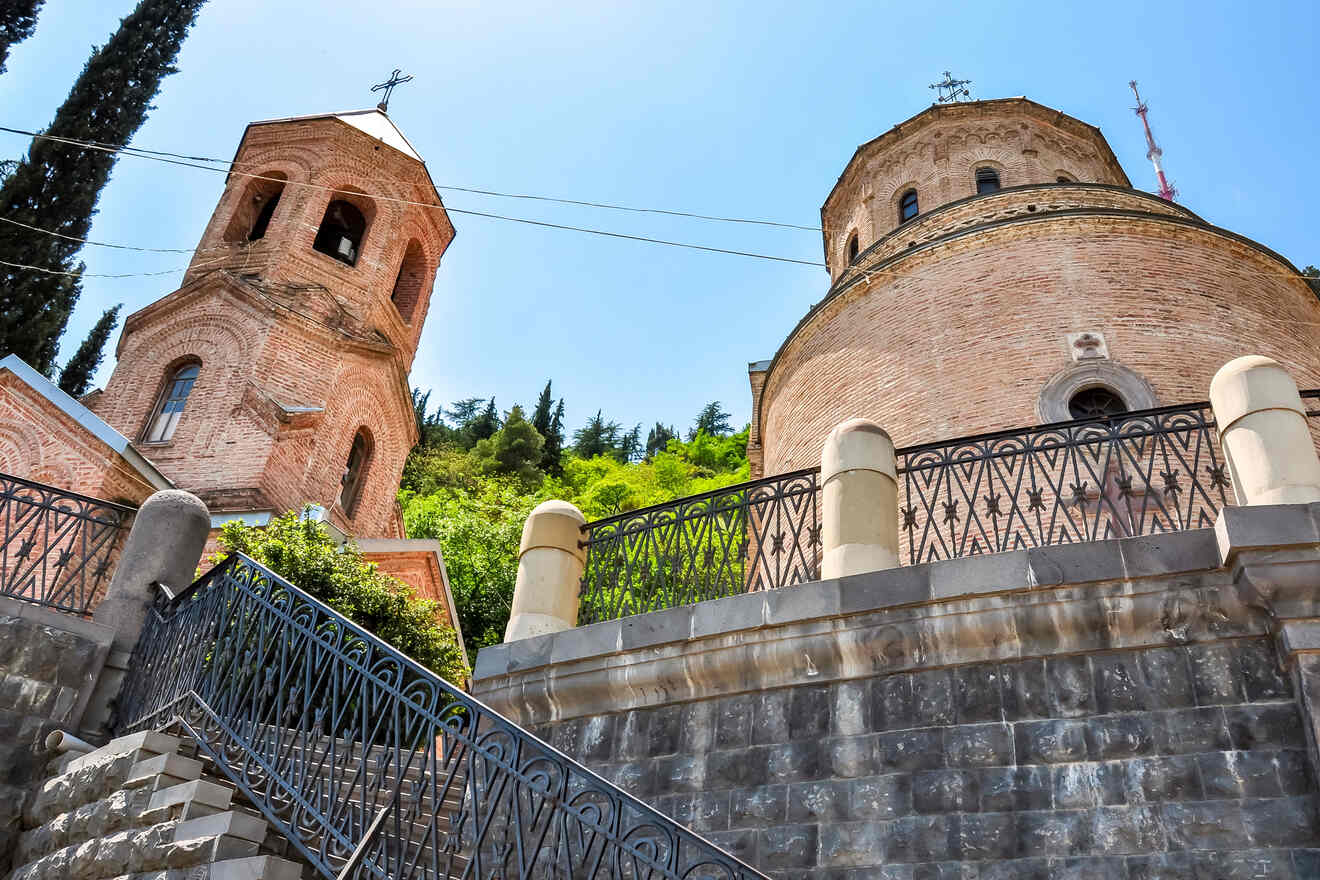 A close-up of the brick-built bell tower of the Mamadaviti Church in Tbilisi, showcasing Georgian ecclesiastical architecture against a backdrop of lush greenery