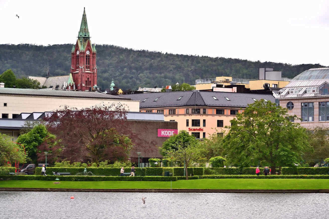 The Johanniskirken spire rises prominently above the cityscape in Bergen, Norway, with the foreground featuring the lush Byparken and the calm waters of Lille Lungegårdsvannet
