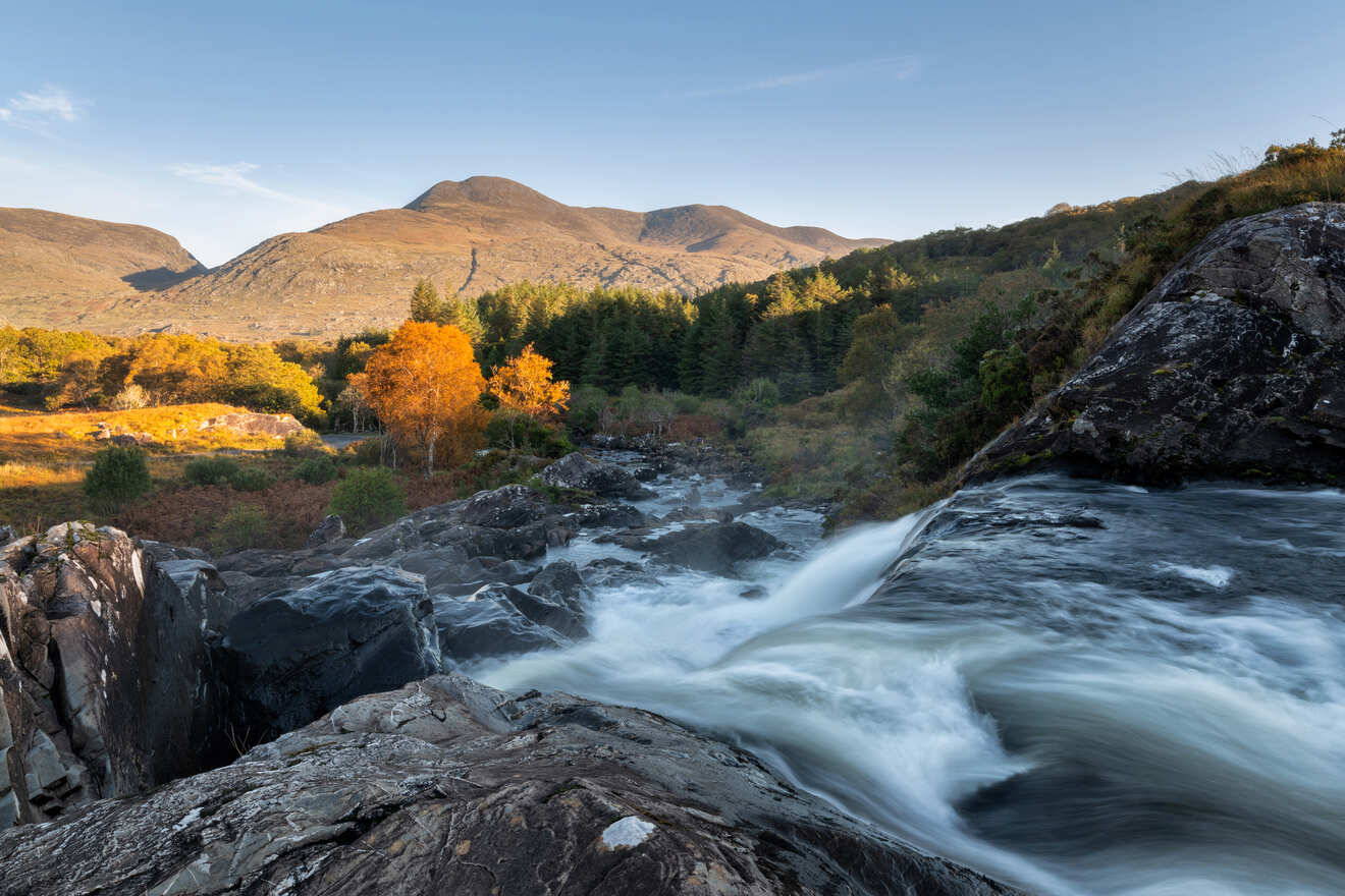 Rushing waterfall with smooth water flow over rocks, surrounded by autumn-colored trees and a backdrop of mountains