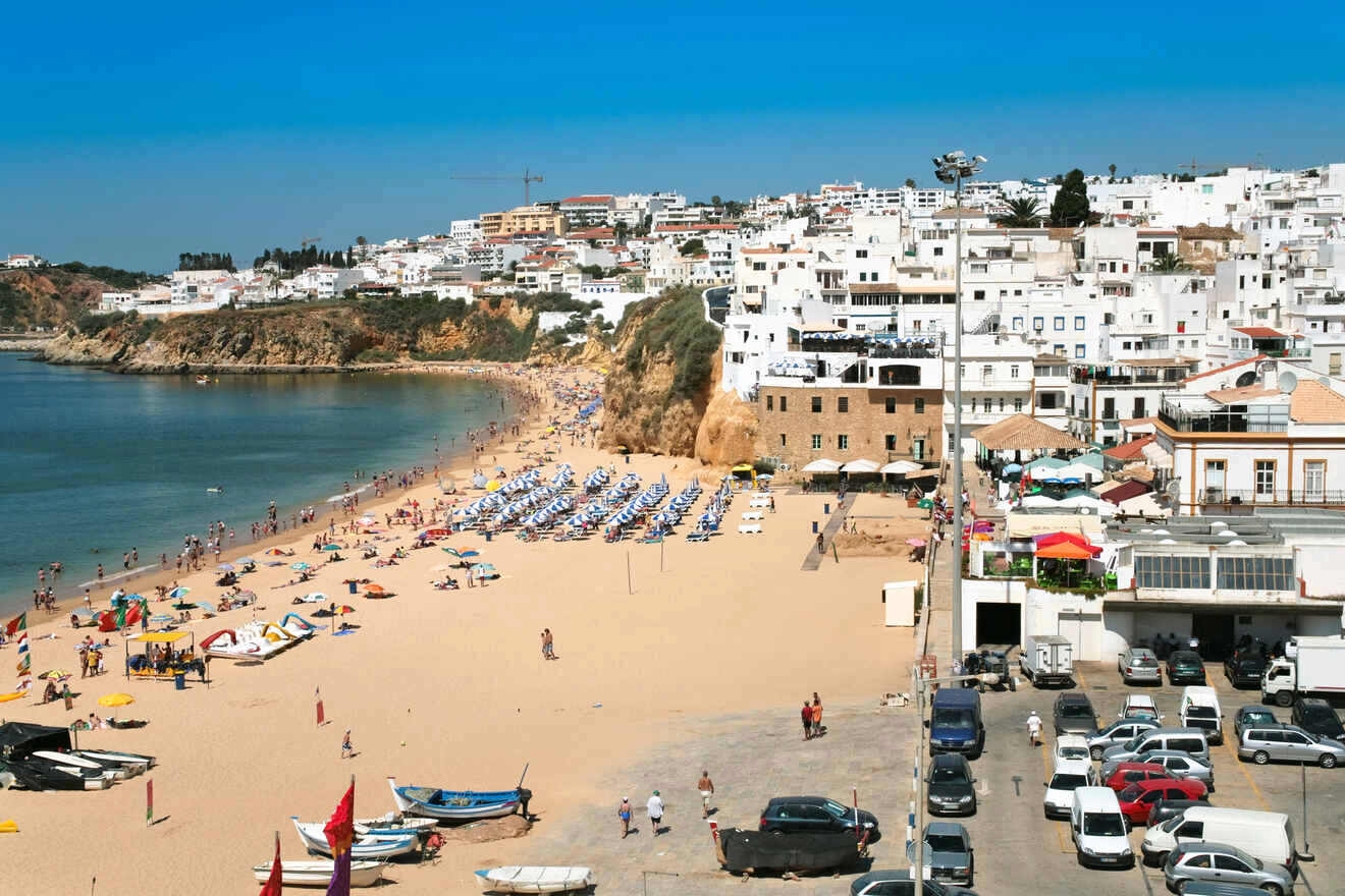 Sandy beach packed with colorful umbrellas and sunbathers, adjacent to a parking area and a picturesque hillside town with white buildings cascading towards the sea