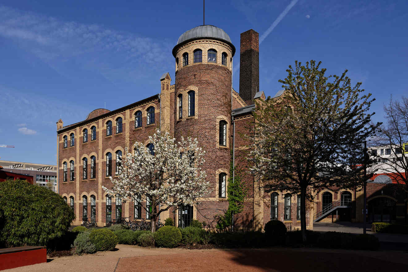 A historic red-brick building with a circular tower, set against a clear sky with blossoming trees in the foreground.