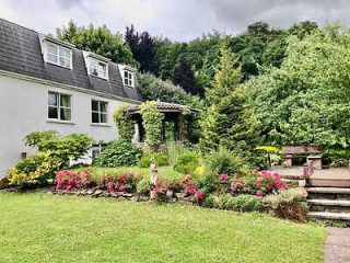 A quaint garden with a lush lawn, pink flowers, and a trellis leading to a white house.