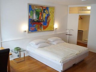 A simple and modern bedroom with a large bed, hardwood floors, abstract artwork above the bed, and soft lighting.