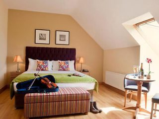 Attic bedroom with a slanted window, green bedspread, colorful pillows, and a violin on a striped ottoman, conveying a cozy and artistic ambiance.