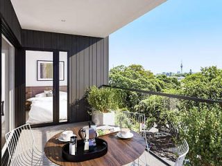 A chic balcony with a round table set for two, offering a sneak peek into a stylish bedroom, and overlooking a lush garden with a view of a city skyline in the distance