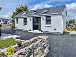 Contemporary white cottage with a blue front door and window shutters, landscaped with a stone wall and young plants.