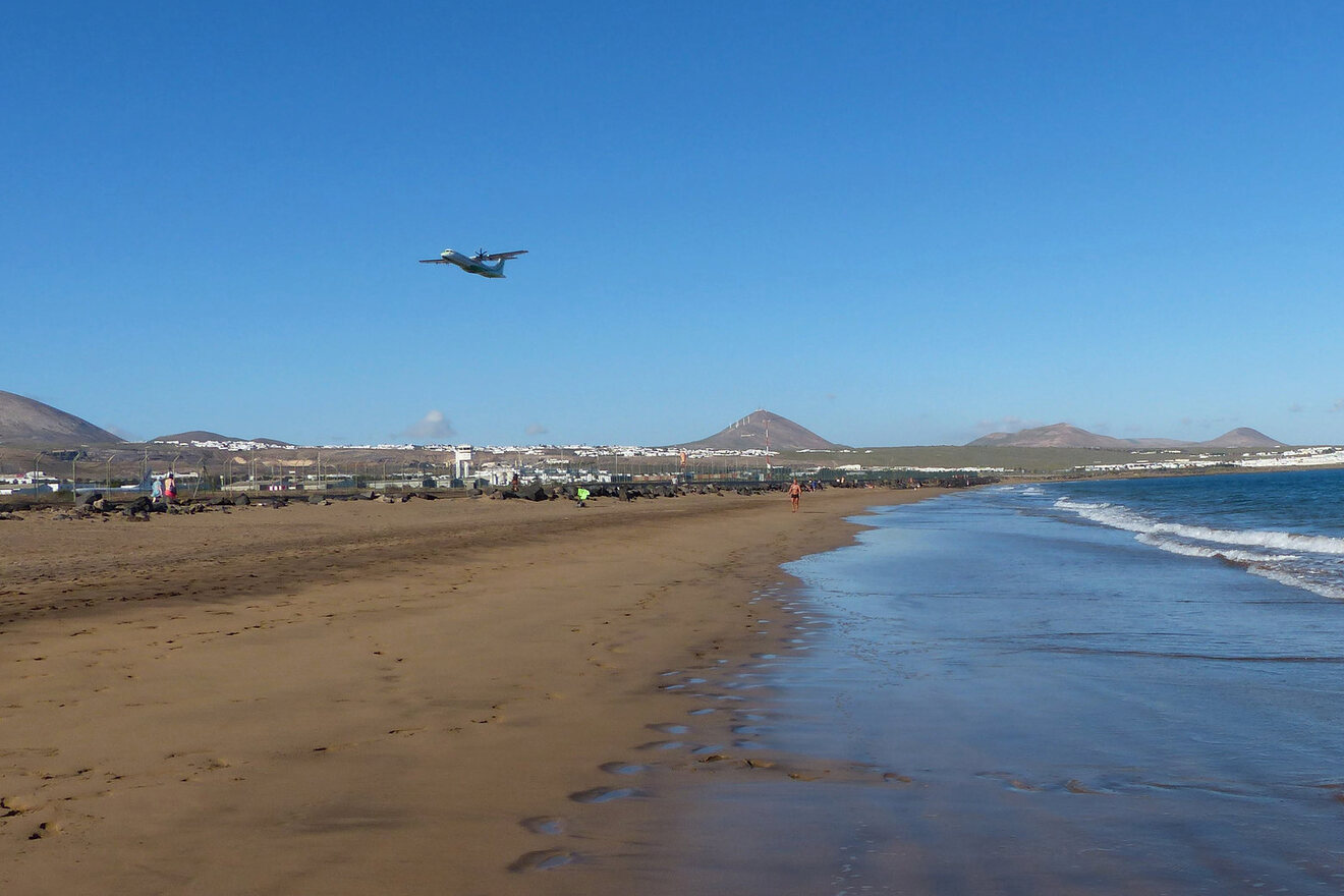 Sandy beach in Lanzarote with an airplane flying low for landing, set against a backdrop of mountains and blue skies