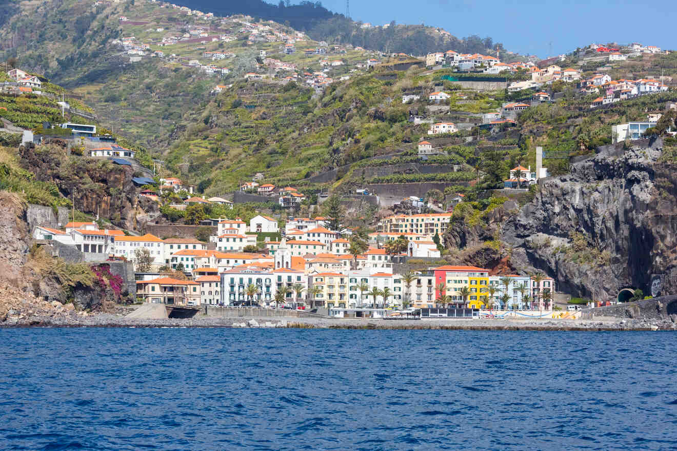 A coastal view from the water, displaying a vibrant town with multicolored buildings and rugged cliffs set against terraced green hills.