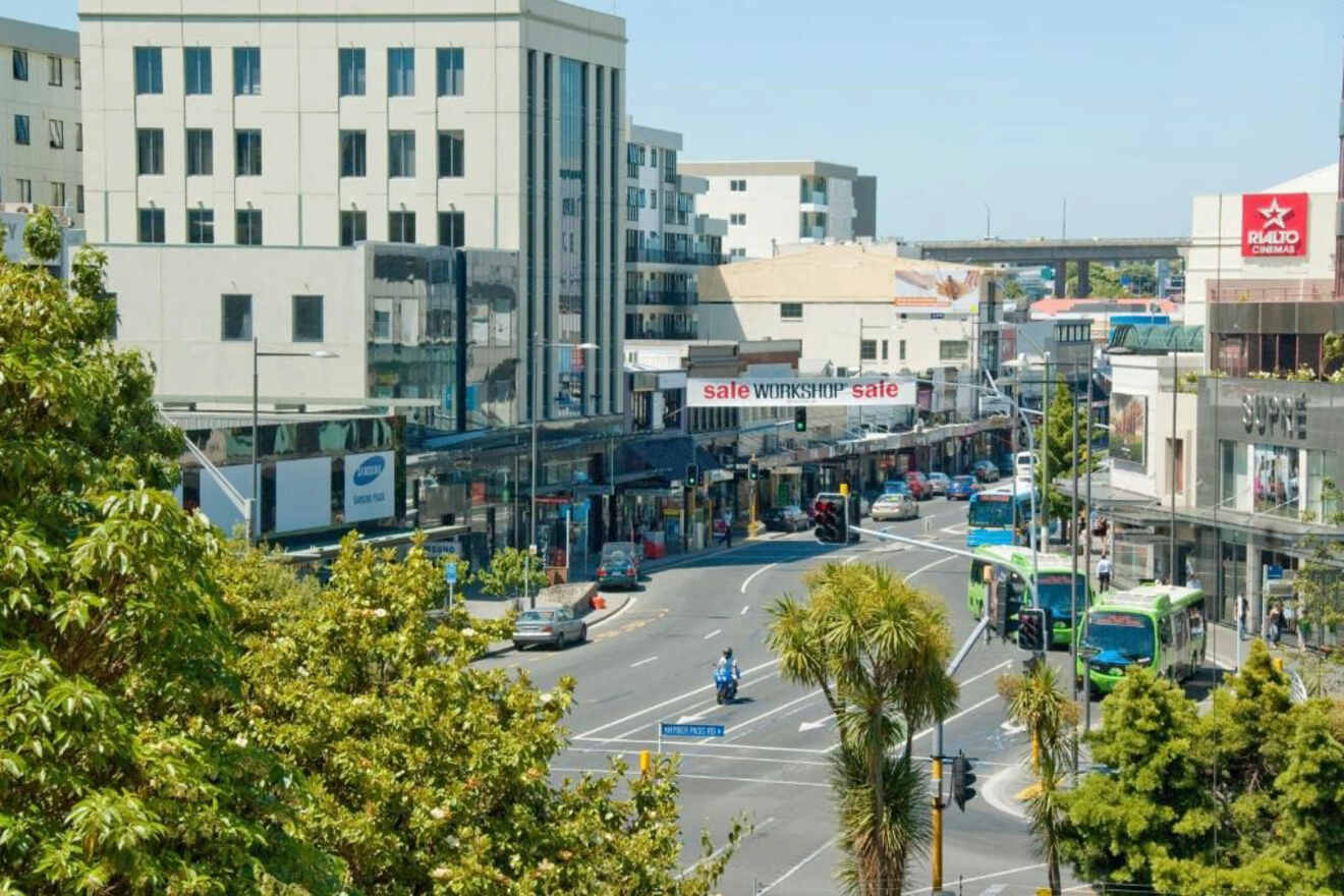 A bustling urban street scene with shops, pedestrians, and public transport, framed by lush trees and a clear sky