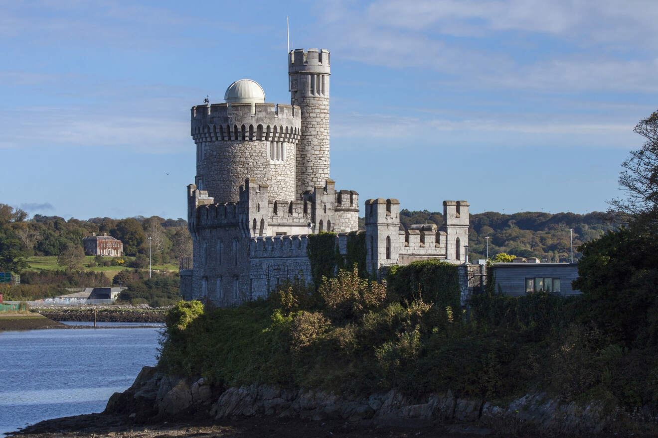 A medieval castle tower in Cork, Ireland overlooking a river, set against a backdrop of greenery and a clear sky