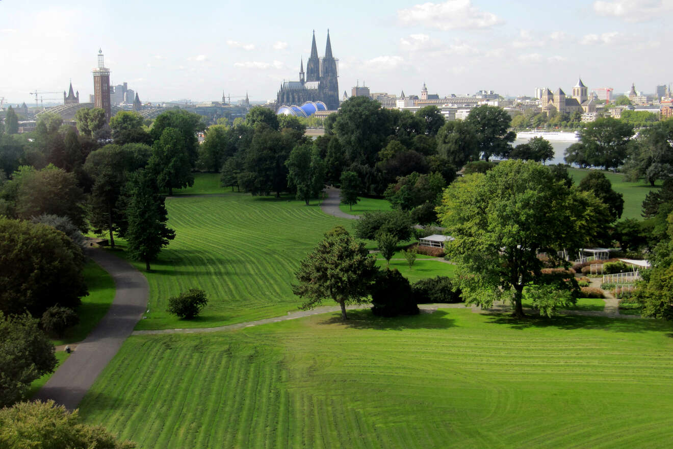 A tranquil park in Cologne with lush green lawns and walking paths, the Cologne Cathedral visible in the distance.