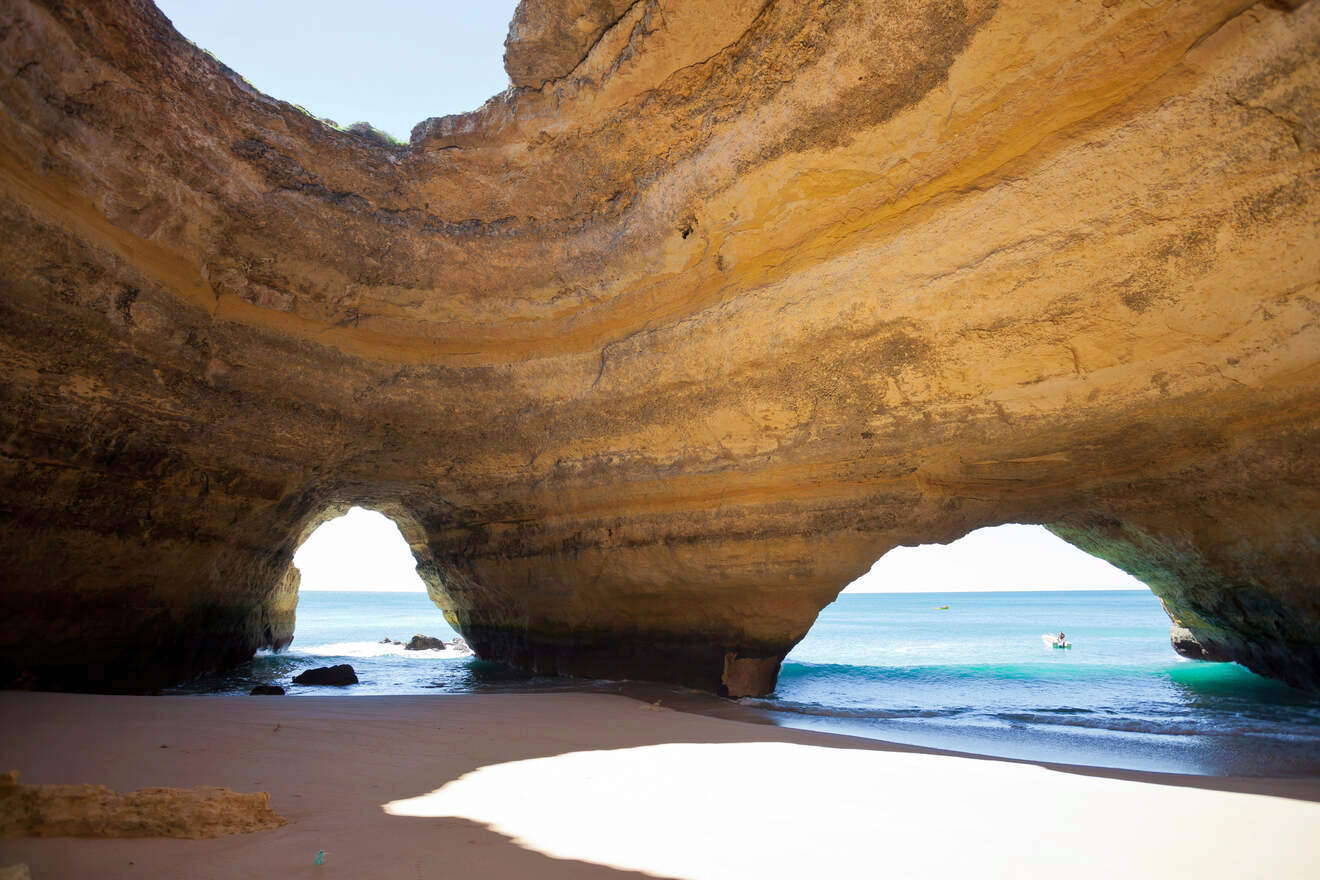 Interior of a natural cave with openings to the sea, showing a sandy beach and clear waters visible through the rock arches