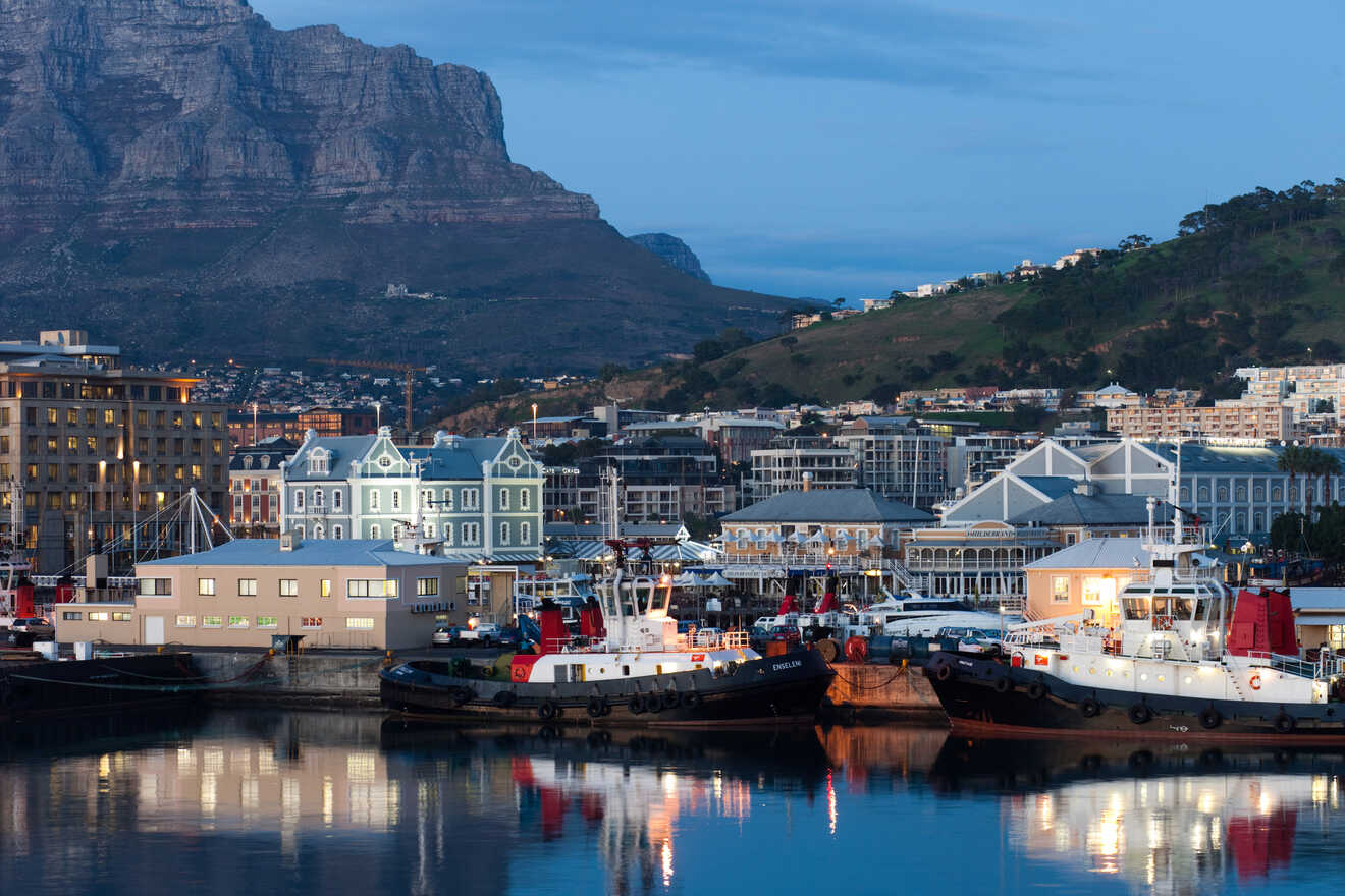 Tranquil harbor scene in Cape Town at dusk with moored boats, waterfront buildings, and a mountain backdrop.