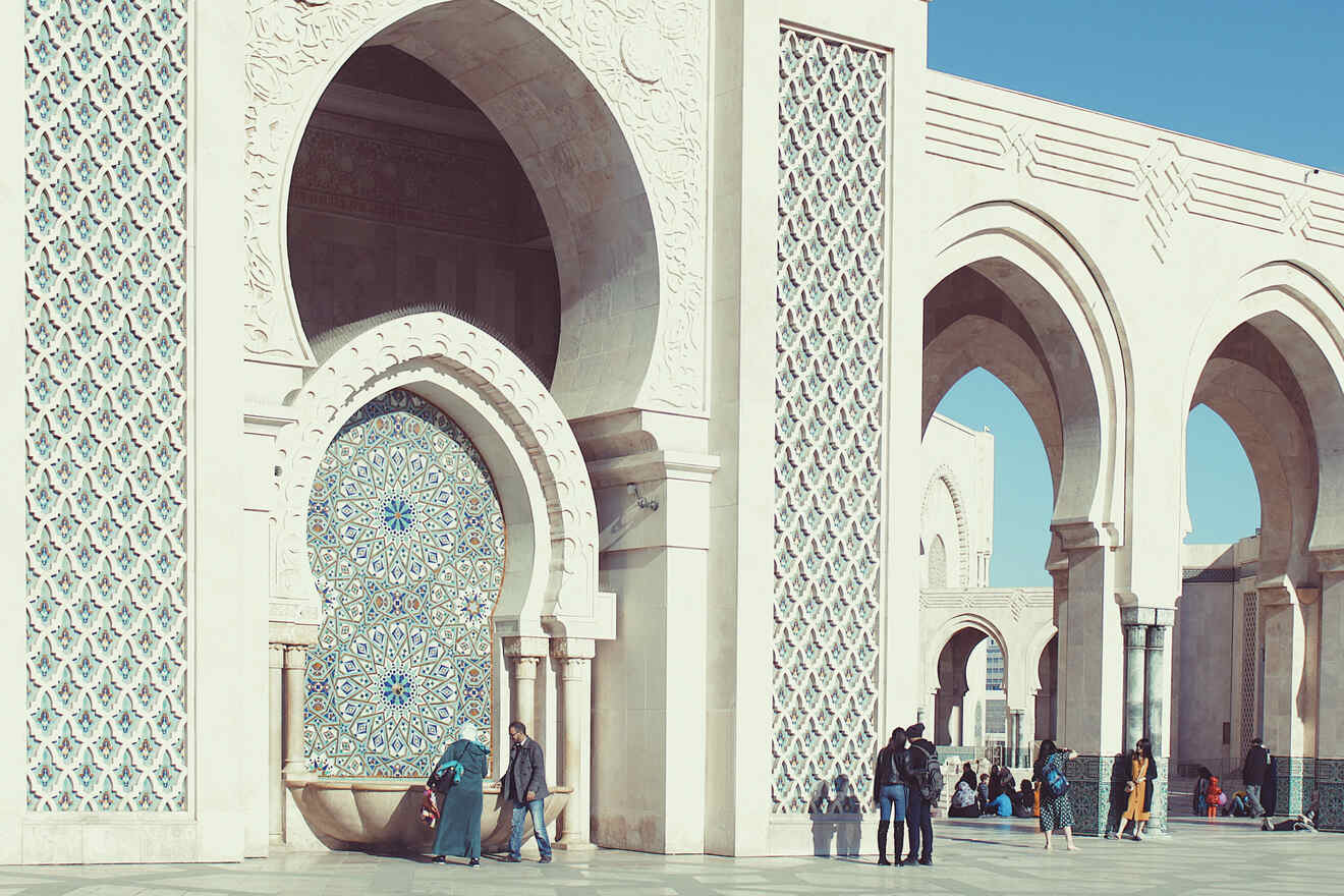 An ornate fountain with intricate blue and white zellij tilework nestled within the arches of a traditional Moroccan architectural style, with people milling about in the sunny square.