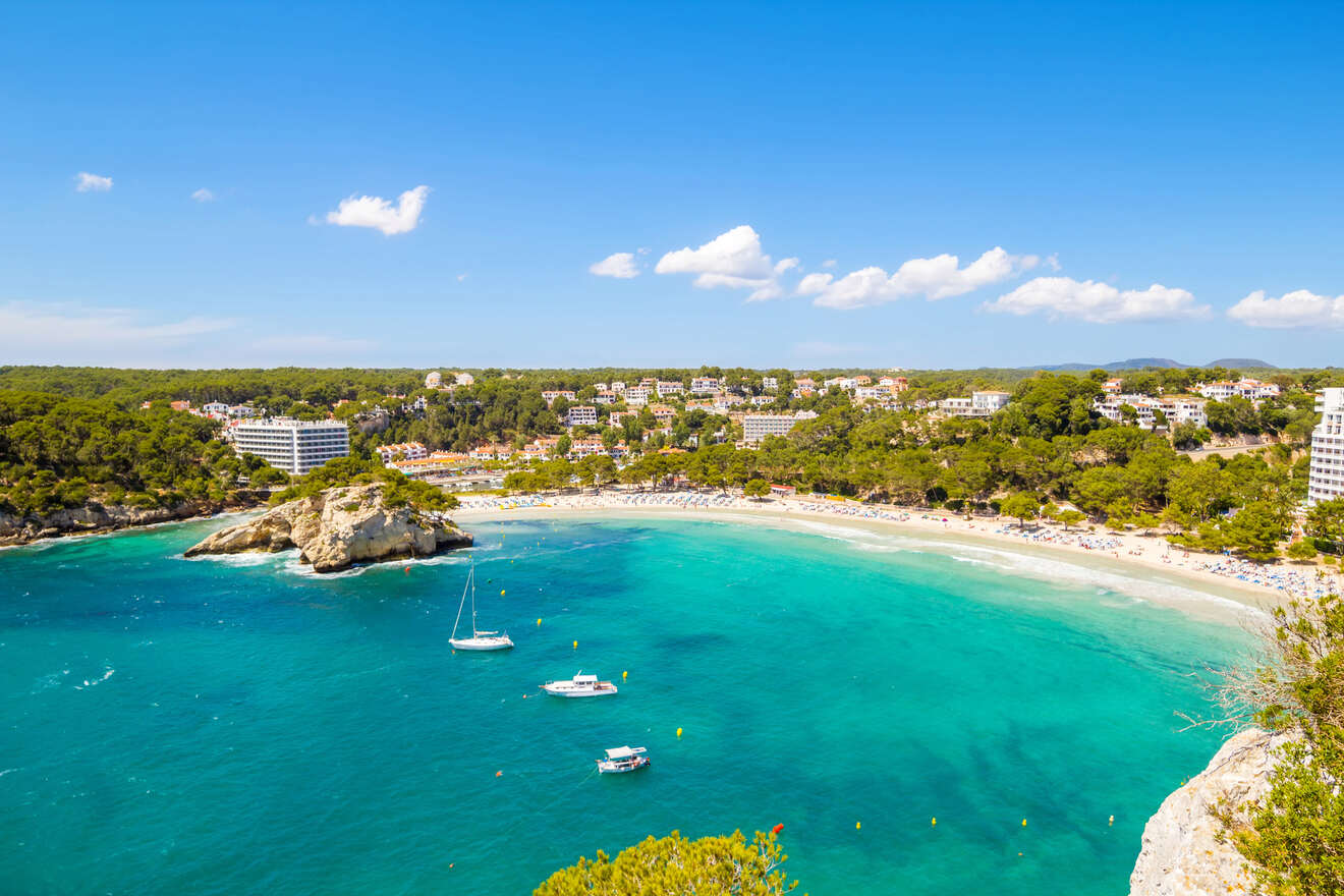 Aerial view of a coastline featuring a crescent-shaped beach with turquoise water, several anchored boats, and a backdrop of trees and buildings under a clear blue sky.