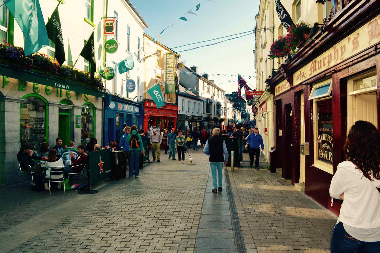 Bustling cobblestone street in a historic town with pedestrians, outdoor cafes, and traditional shop fronts under festive banners