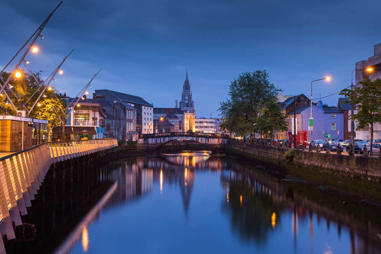 A tranquil evening view of a city riverfront in Cork, Ireland with illuminated streets and buildings, and a cathedral in the distance reflected in the water.
