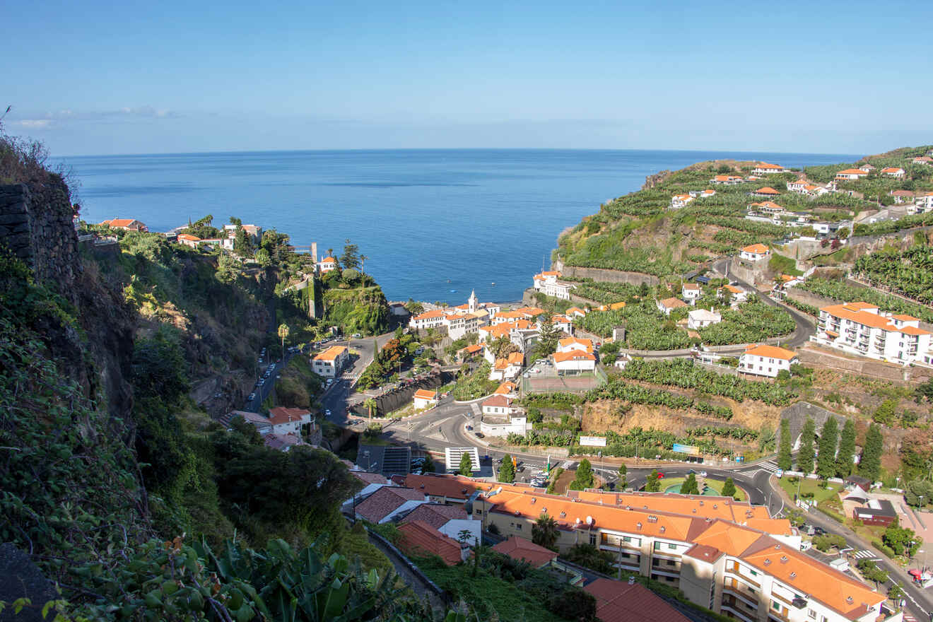 A panoramic view of a coastal town with terraced hills, winding roads, and a deep blue sea stretching to the horizon under a clear sky