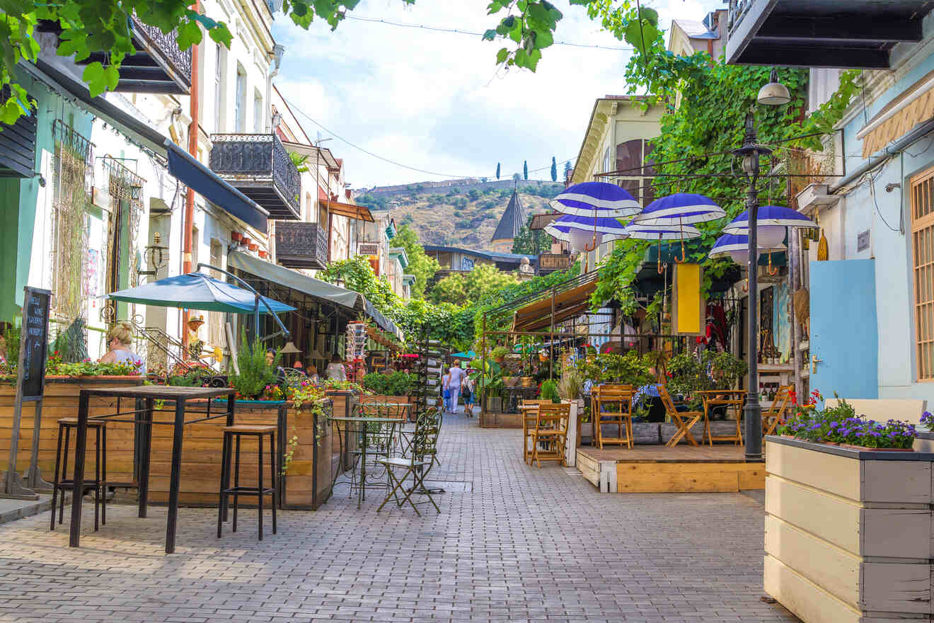 A charming pedestrian street lined with colorful outdoor cafés and bistros, adorned with greenery and flowers under a blue sky, with cobblestone paving leading towards distant hills.