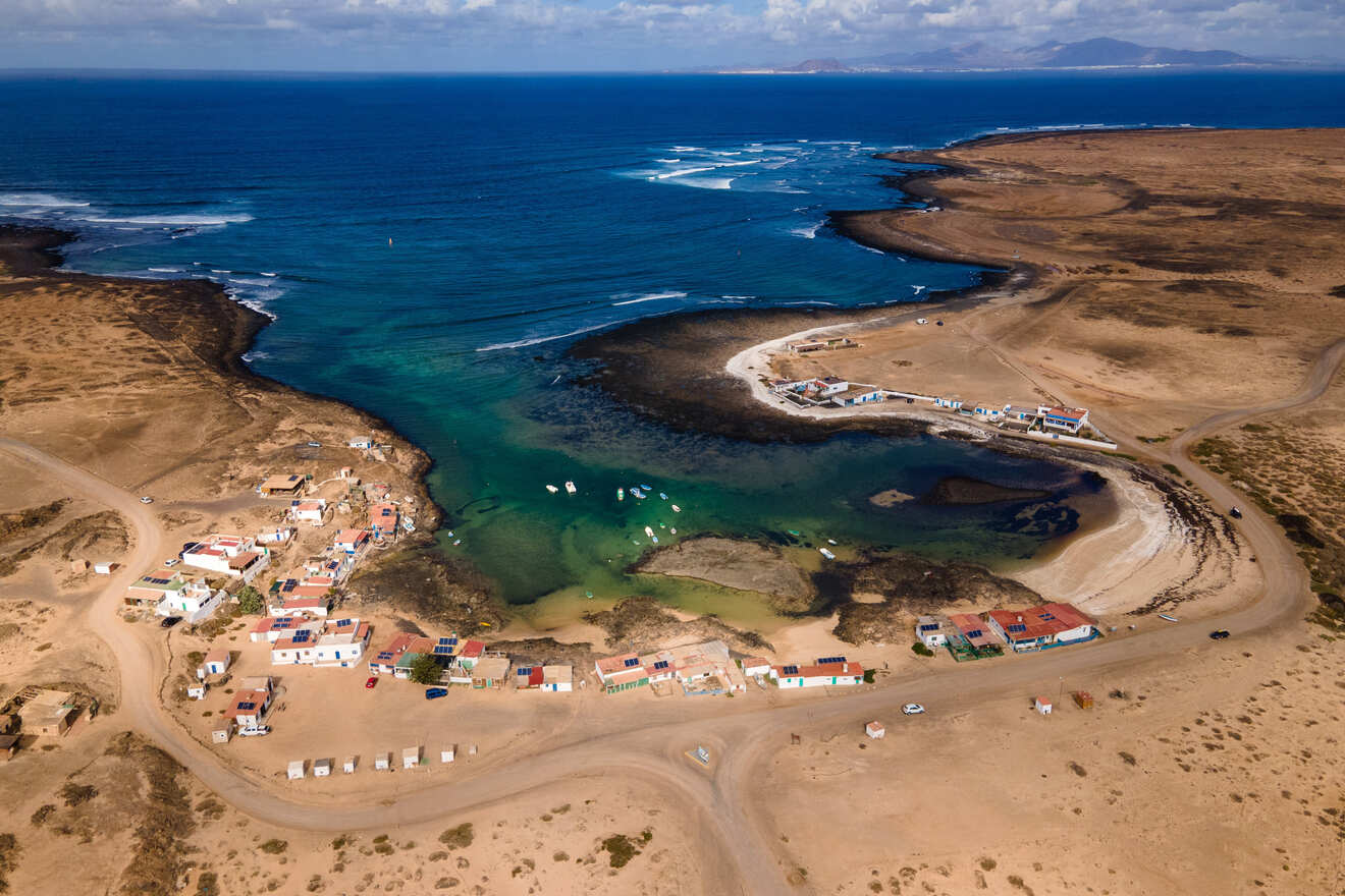 Aerial shot of a tranquil fishing village in Fuerteventura, nestled in a cove with boats moored in calm lagoon waters, flanked by a rugged landscape and the open sea.