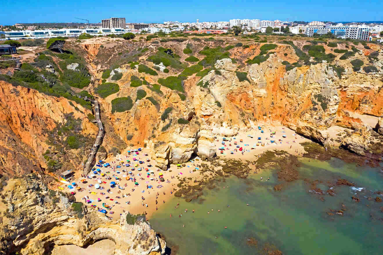 Aerial view of a sun-soaked beach in Algarve nestled between dramatic red cliffs and the urban landscape of a city, with beachgoers enjoying the golden sands