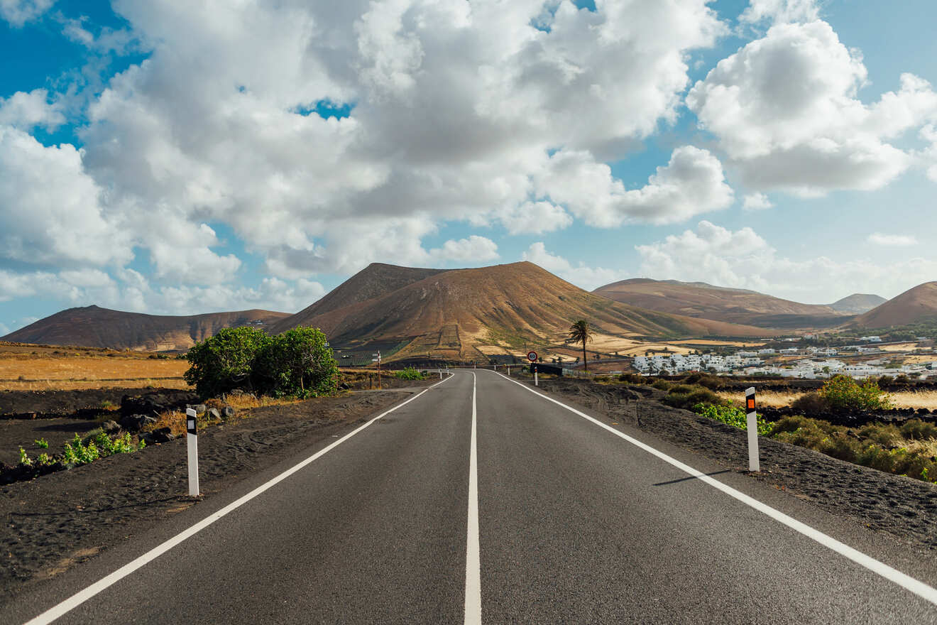 Road leading through the unique landscape of Lanzarote, with volcanic mountains on the horizon and a blue sky with fluffy clouds