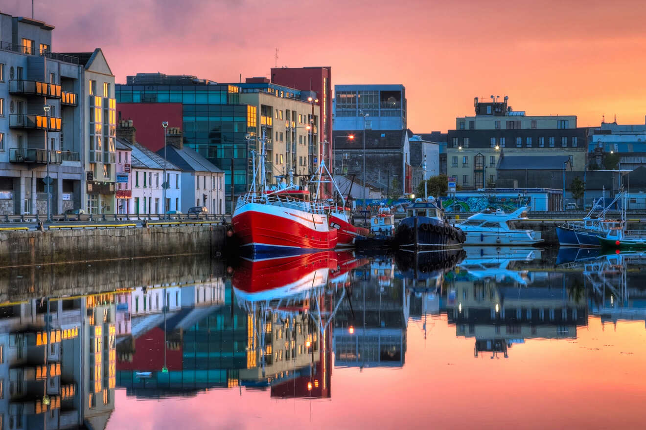 Tranquil harbor at dusk with a red fishing boat, modern buildings, and calm waters reflecting the vibrant sunset sky and city lights, showcasing a serene maritime setting.