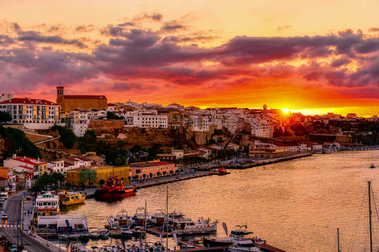 Sunset at Menorca's coast, with the golden hour casting warm hues over the historic architecture of the city, a marina filled with boats, and the open sea, reflecting the vibrant sky