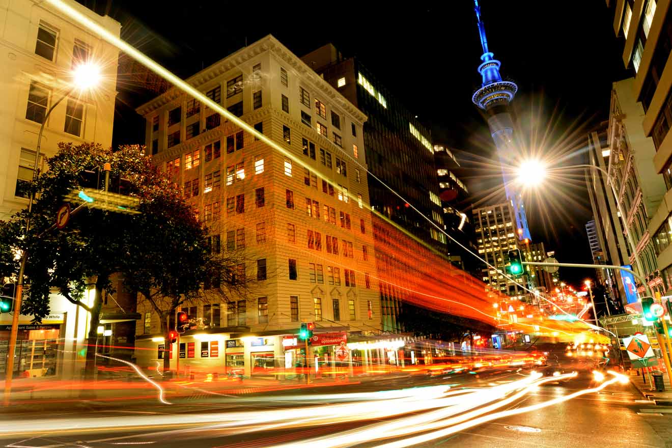 Nighttime city street scene with long-exposure light trails from moving traffic, brightly illuminated buildings, and the Sky Tower in the background