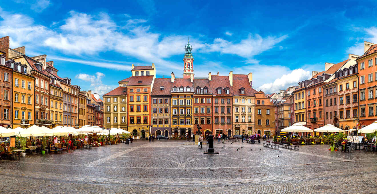 Historic European square with colorful buildings, café seating under umbrellas, cobblestone pavement, and a central statue.