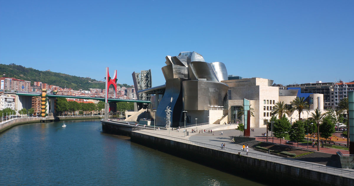 The Guggenheim Museum Bilbao, a modern art museum with a distinctive metallic exterior, stands alongside the Nervión River in Bilbao, Spain, under a clear blue sky.