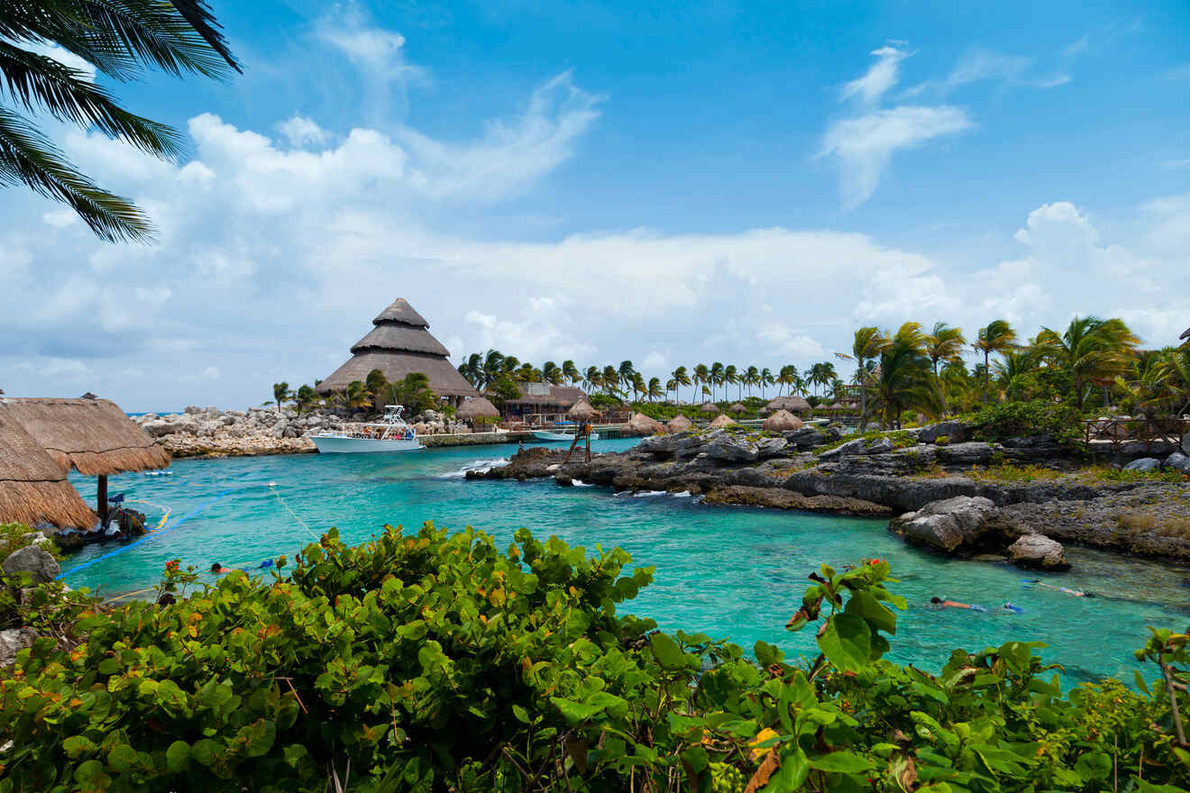 Tropical paradise at Xcaret Park, featuring traditional thatched structures amidst clear blue waters, rocky formations, and lush greenery, with visitors enjoying the aquatic activities