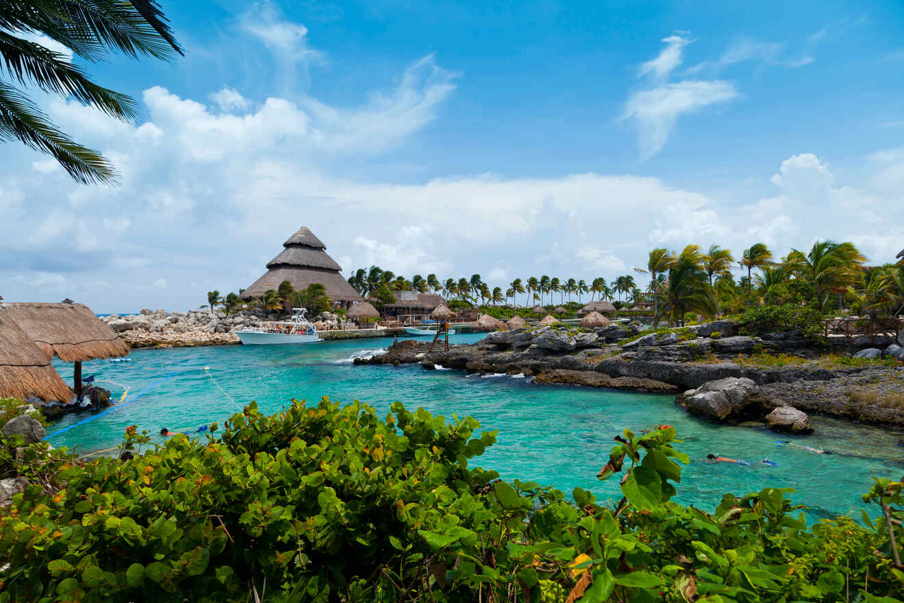 Scenic view of a tropical resort with crystal-clear turquoise water, rocky shoreline, lush greenery, and thatched-roof buildings under a partly cloudy sky.