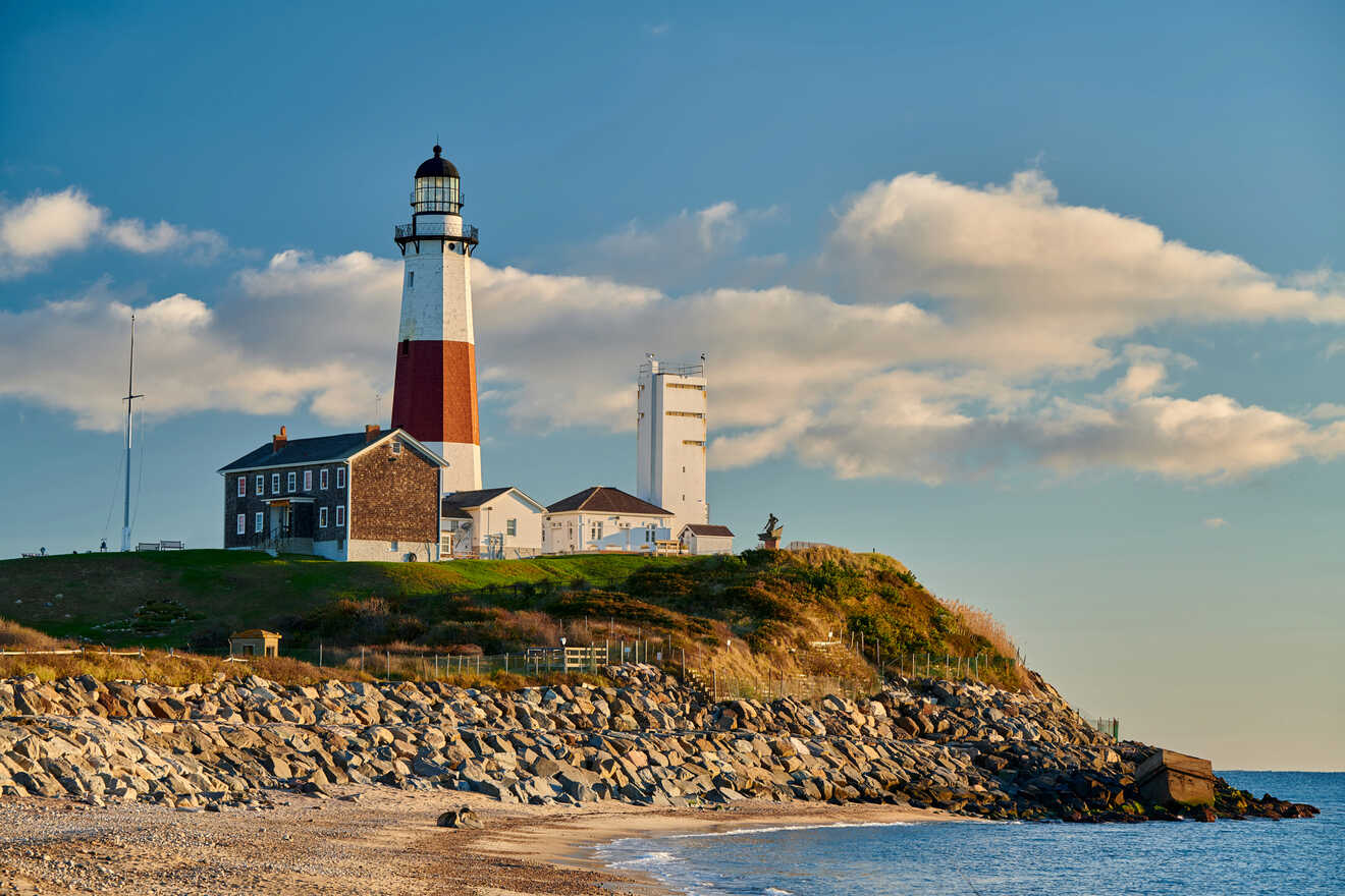 A historic lighthouse in the Hamptons with red and white stripes stands on a grassy cliff, overlooking a rocky shore and calm sea under a blue sky with soft clouds