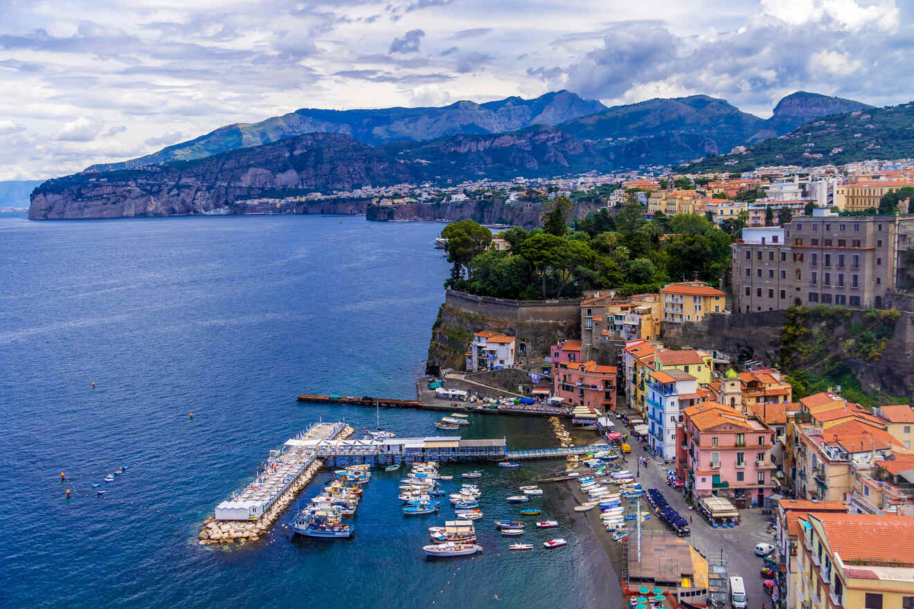 Coastal view of Sorrento showing multi-colored buildings, a marina with boats, and cliffs with a backdrop of mountains under a cloudy sky