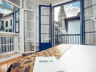 Hotel room with open balcony doors revealing a view of a historic church spire, and a notepad with 'Sawday’s' branding on the bed.