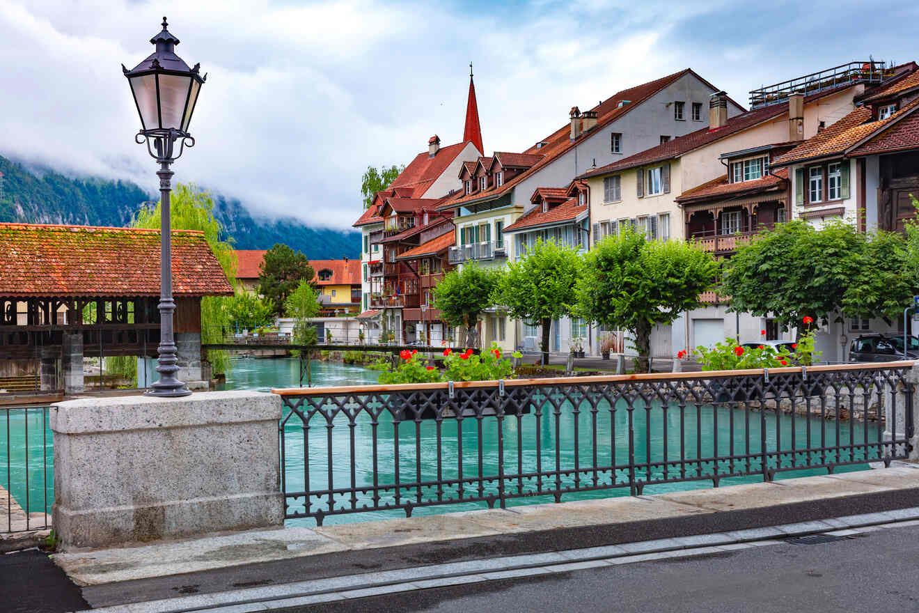 Quaint riverside scene in Interlaken with traditional Swiss houses, blooming flowers, and a covered wooden bridge, set against a backdrop of misty mountains