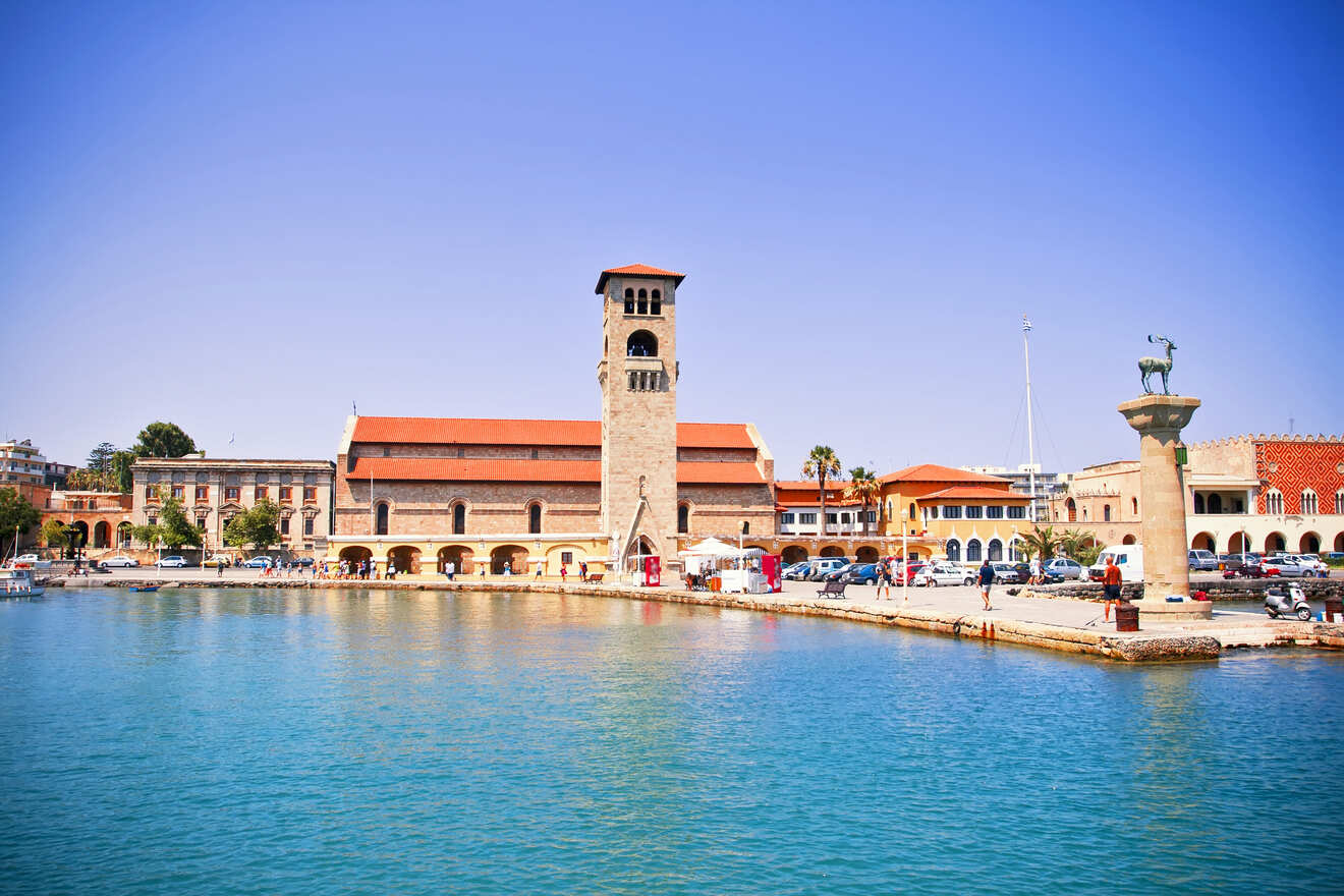 A waterfront view of a church tower in Rhodes, with surrounding buildings and a statue of a deer near the marina