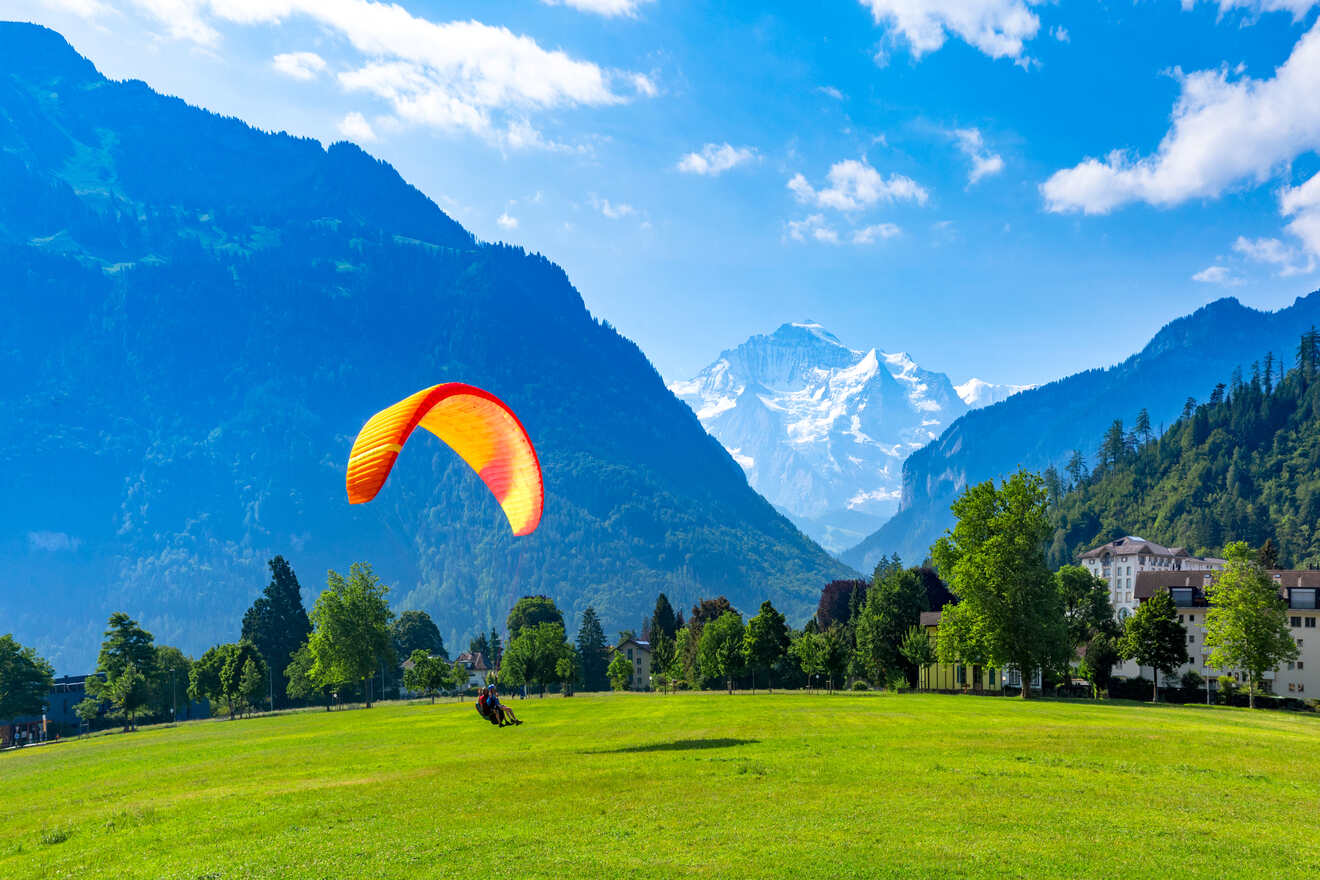 Paragliding over a lush green field in Interlaken with the Swiss Alps towering in the background and a clear blue sky above
