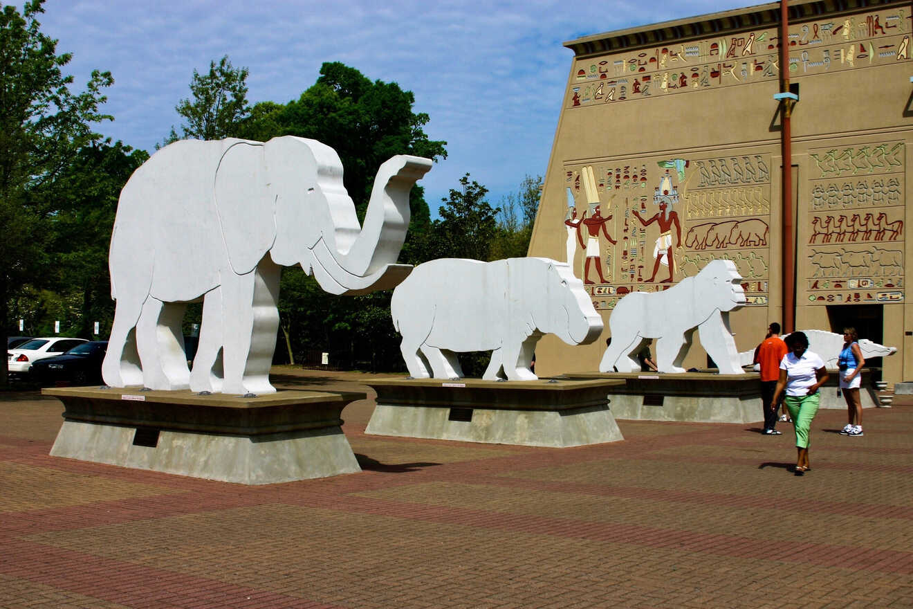 A series of large white animal sculptures, including elephants and lions, in front of the Memphis Pyramid, an iconic landmark, with visitors walking around the plaza area