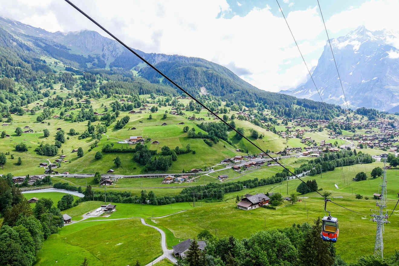 Aerial view of a cable car ascending over the green valley of Interlaken, with alpine houses dotted across the landscape and mountains in the background