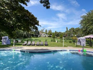 An outdoor swimming pool with a curved design, lined with multiple sunbeds and umbrellas, set in a well-maintained lawn with trees in the background