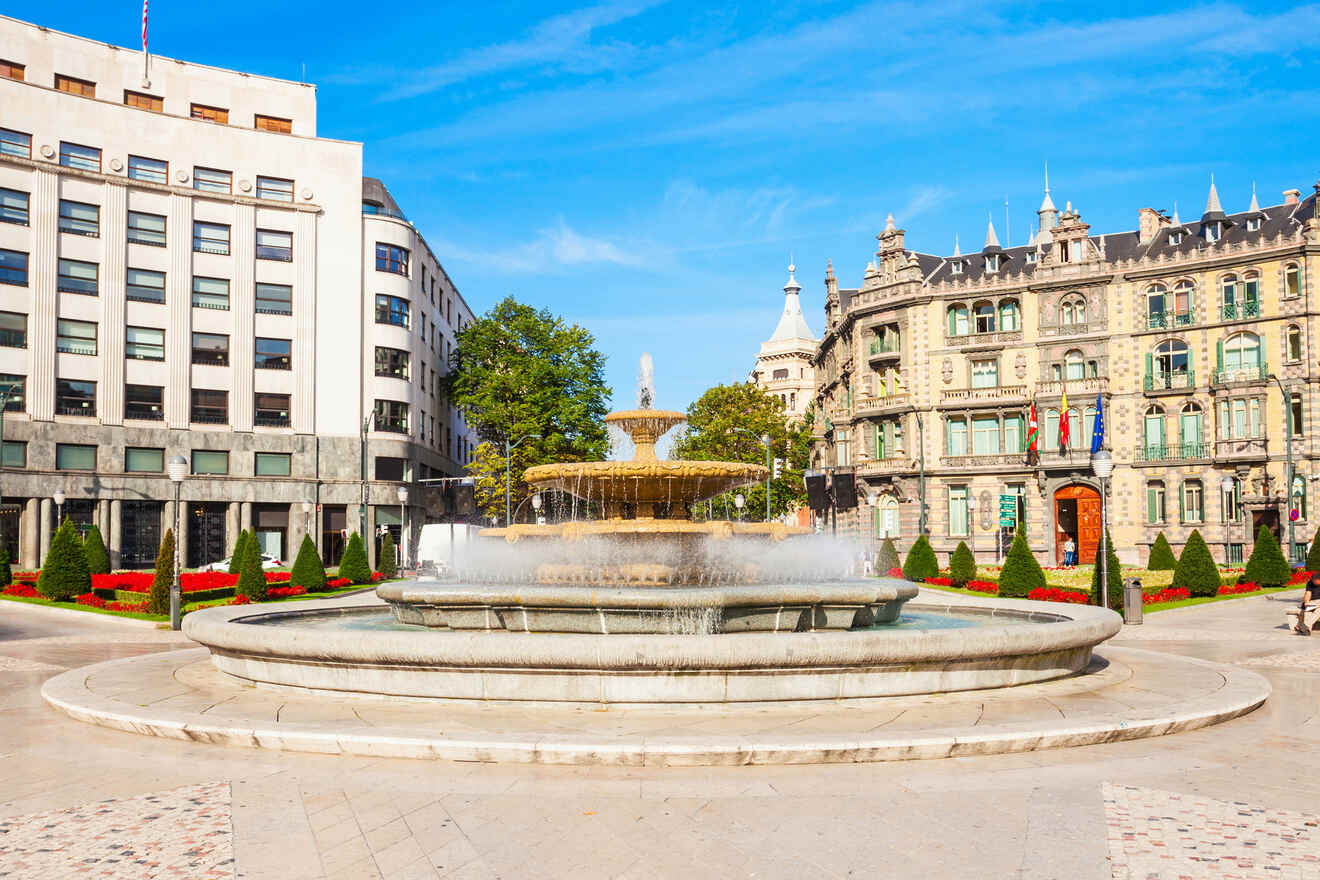 City square in Bilbao with an ornate fountain, modern and classical buildings, and vibrant flower beds under a clear sky.