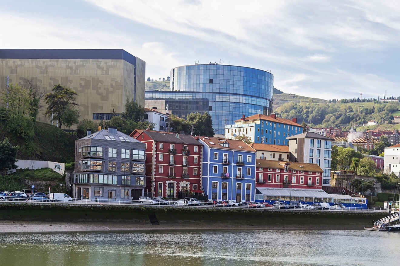 Contrast of old and new architecture along a riverbank, with colorful traditional houses in the foreground and modern glass buildings in the background, under a hilly landscape