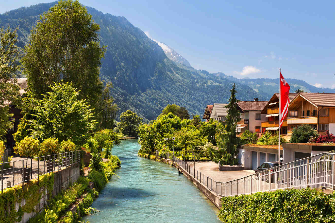 Idyllic riverside walkway in Interlaken flanked by lush greenery, Swiss flags, and a clear view of the Alps in the distance